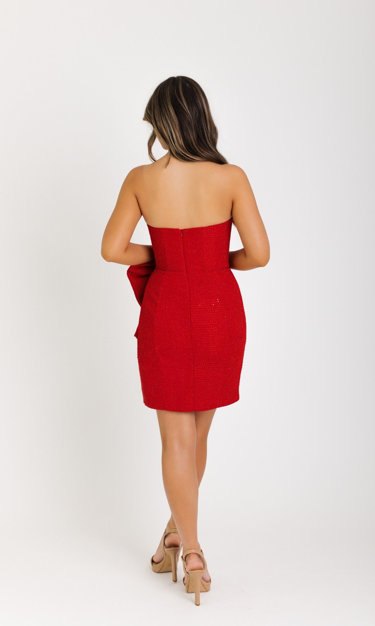 A woman stands facing away in the Ava Presley 42917 short homecoming dress, a strapless red design with a statement bow, paired with beige heels against a plain white background.